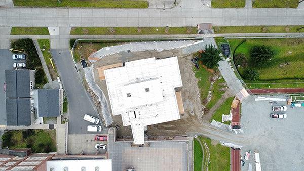 aerial view directly over a building under construction showing a narrow connection to the existing building, a road at the top of the image, and a parking lot to the left